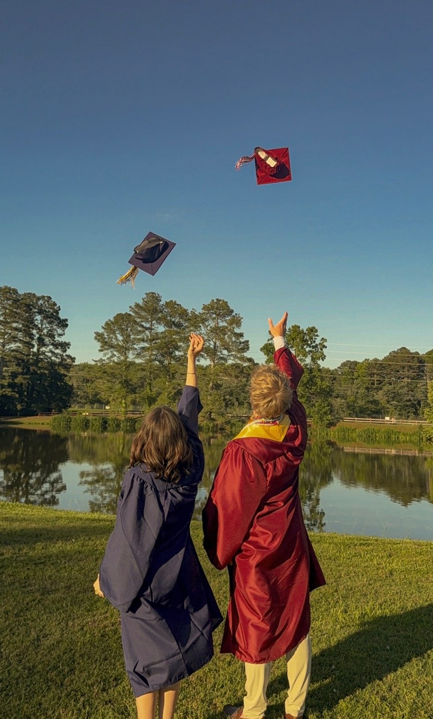Two graduates in caps and gowns tossing their caps in the air beside a lake